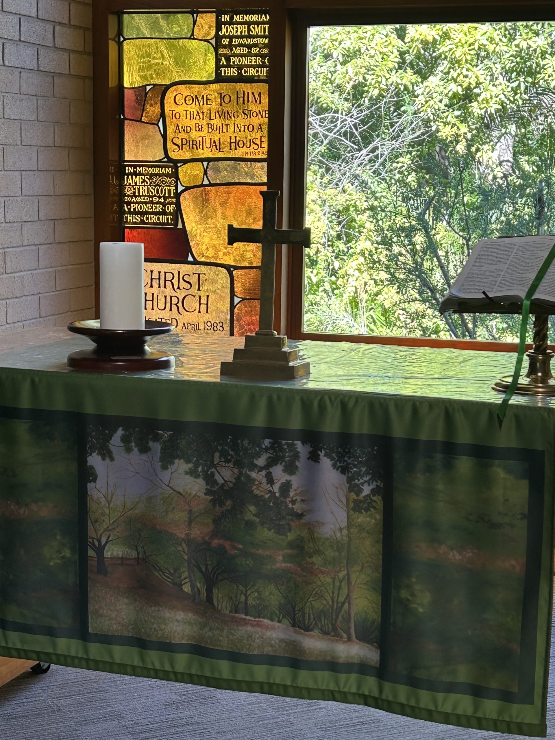 Communion Table, green cloth, gold cross, bible and white candle, in front of stained glass window with view to trees